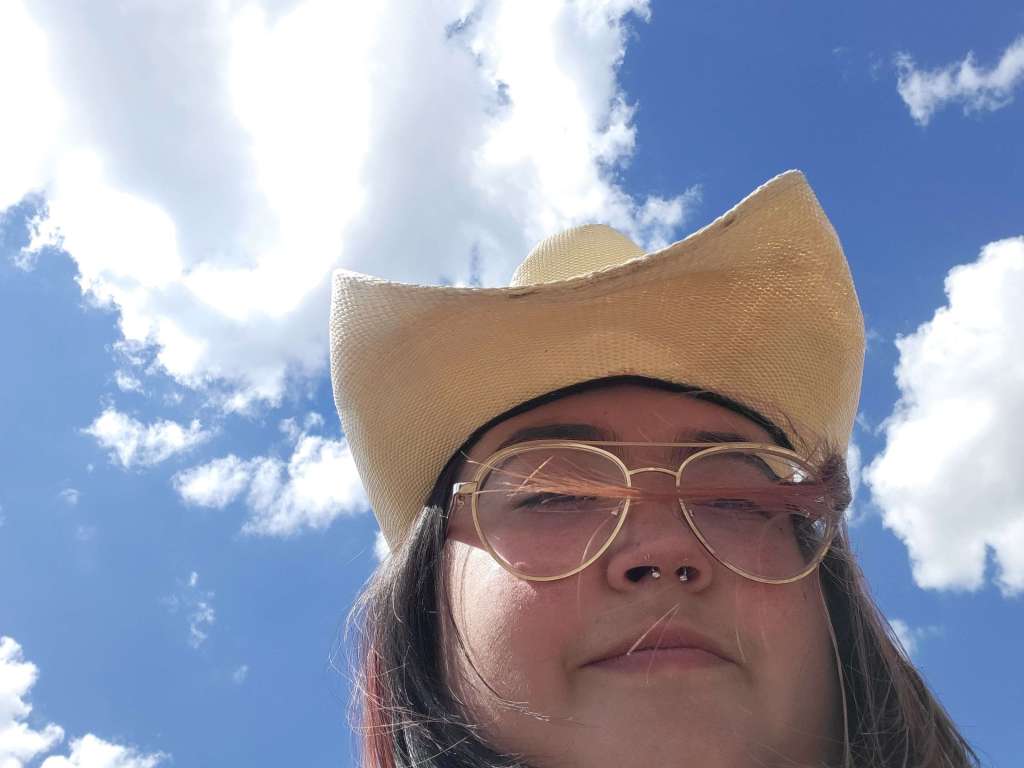 SG Huerta facing down at the camera wearing a vaquero hat. the background is a blue sky with fluffy clouds